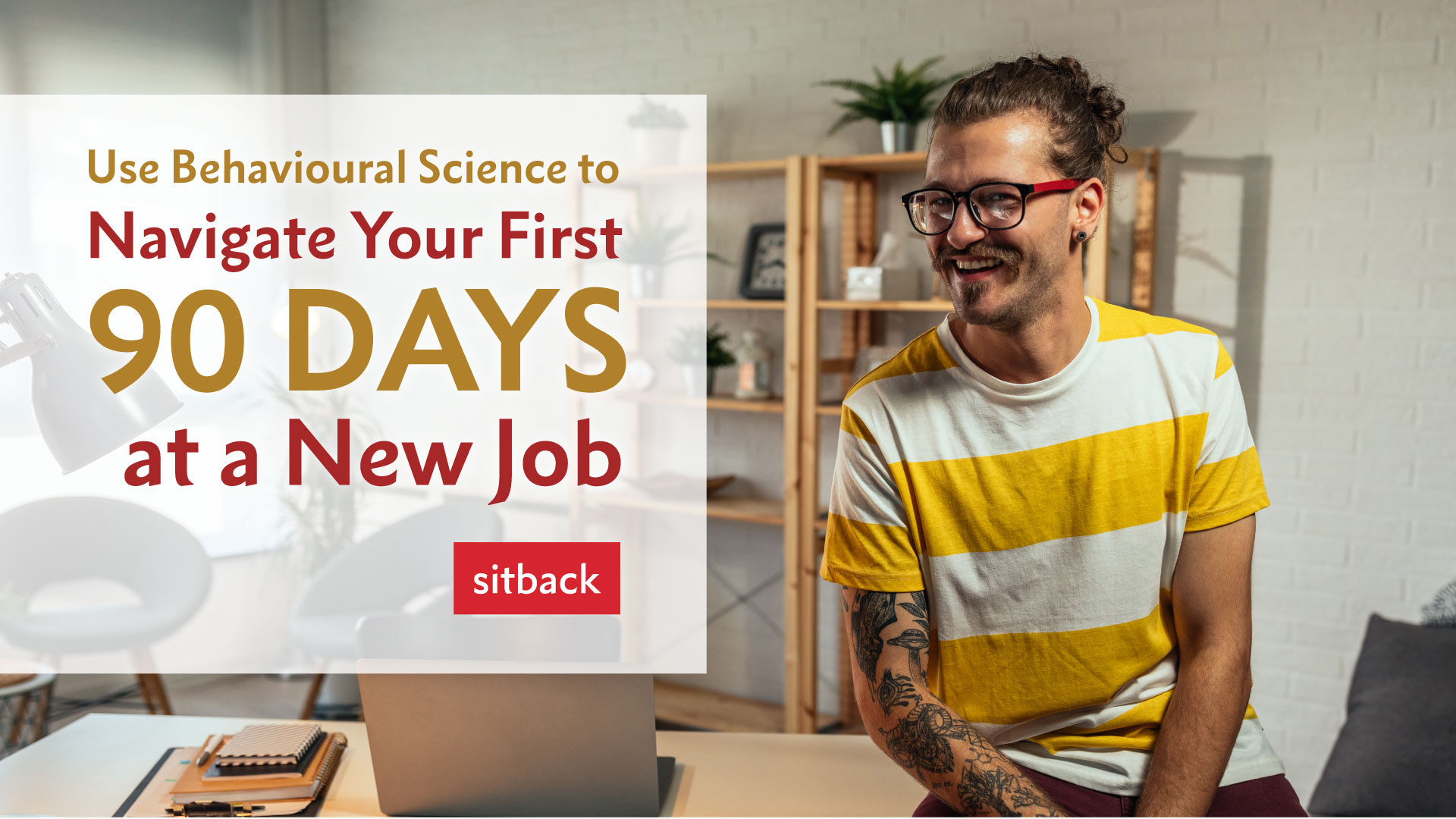 Man in glasses wearing a stripy t-shirt, sat on top of a desk and smiling as he starts his new job because he knows that he is using our behavioural science-backed tips to ensure success